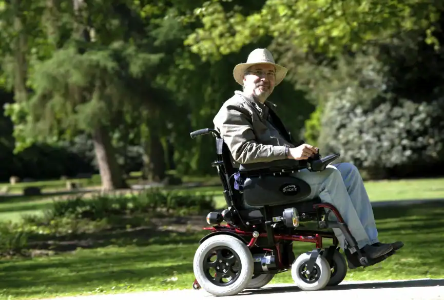 A man wearing a hat and glasses rides a durable medical equipment electric wheelchair on a paved path through a park with green trees and grass.