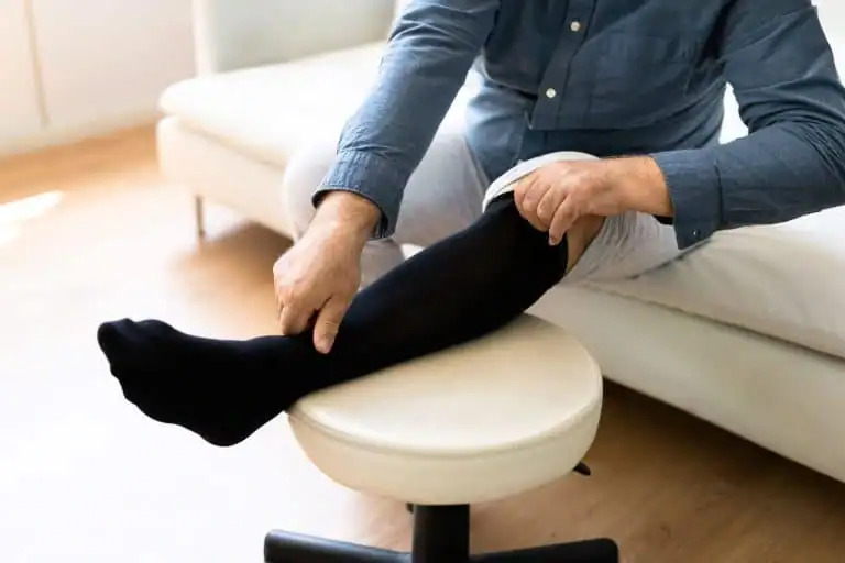 A person sitting on a couch puts on a black compression stocking, an essential consumable medical supply, with their foot resting on a round stool.