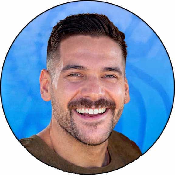 A man with short dark hair, trimmed beard, and mustache smiles at the camera in front of a blue background, reminiscent of a portrait style often seen in Kristin Hoffman's photography.