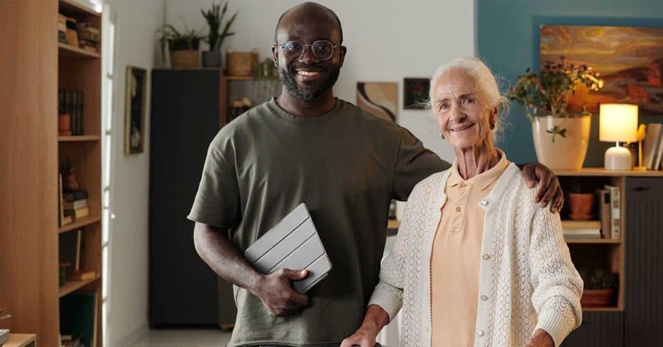 A smiling man holding a tablet stands with his arm around an elderly woman in a bright living room, highlighting the importance of home safety and dementia safety in everyday life.