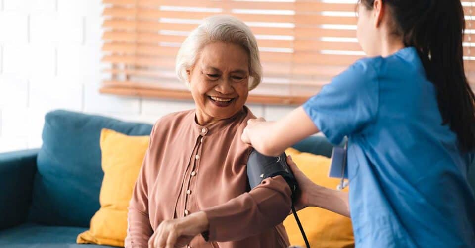 A nurse in blue scrubs checks the blood pressure of a smiling elderly woman sitting on a couch with yellow pillows, making sure to review the Discharge Equipment Checklist for her safe transition home.