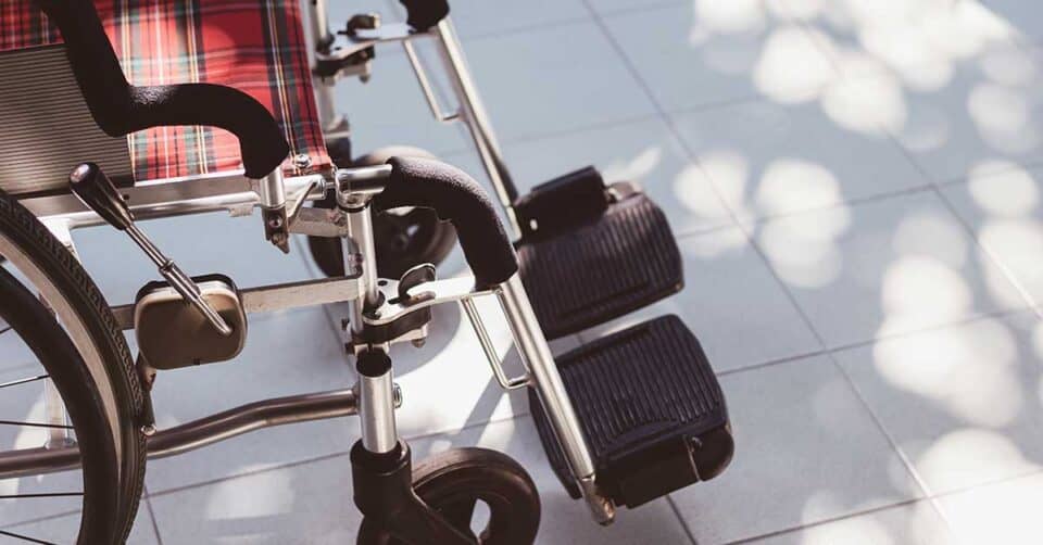 A close-up view of an empty wheelchair with a plaid seat, positioned on a tiled floor with dappled sunlight and shadows—an essential piece of home medical equipment designed for comfort and mobility.