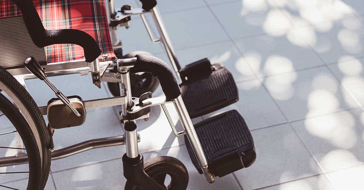 A close-up view of an empty wheelchair with a plaid seat, positioned on a tiled floor with dappled sunlight and shadows—an essential piece of home medical equipment designed for comfort and mobility.