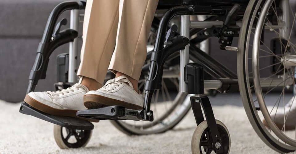 Close-up of a person’s legs in beige pants and white sneakers, seated in a wheelchair on a light-colored carpet—an important reminder of the role mobility aid maintenance plays in everyday comfort and ease.