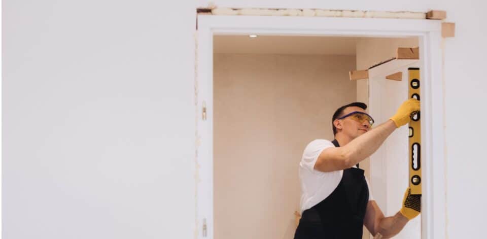 A worker wearing safety glasses and yellow gloves uses a level to check the alignment of a door frame for a wheelchair accessible home.