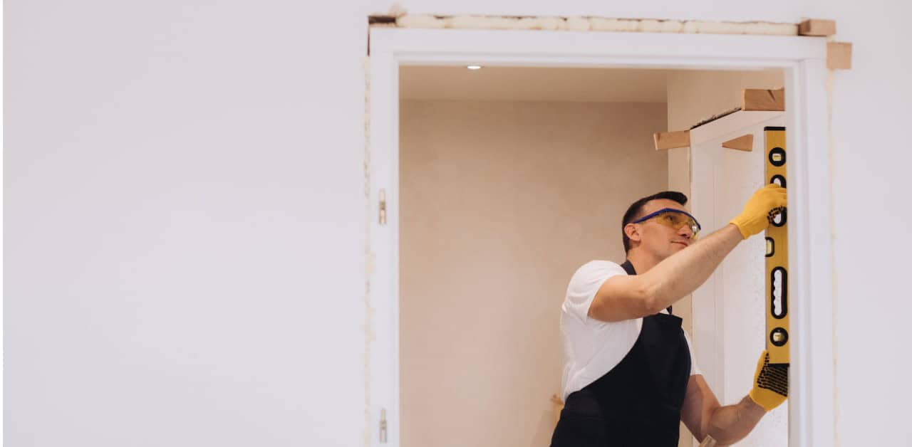 A worker wearing safety glasses and yellow gloves uses a level to check the alignment of a door frame for a wheelchair accessible home.