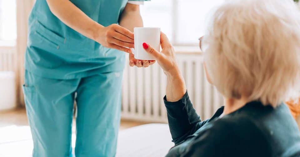 A healthcare worker in blue scrubs hands a white mug, possibly containing nutritional supplements, to an older woman with gray hair, who is sitting down indoors.