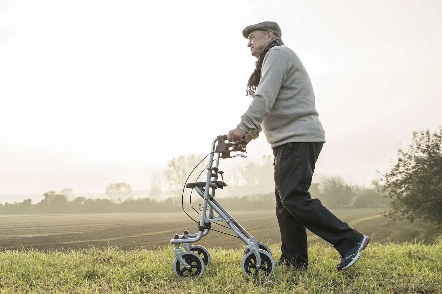 An older man wearing a hat and scarf uses durable medical equipment—a walker—while standing on grassy ground near a field, with a hazy sky in the background.