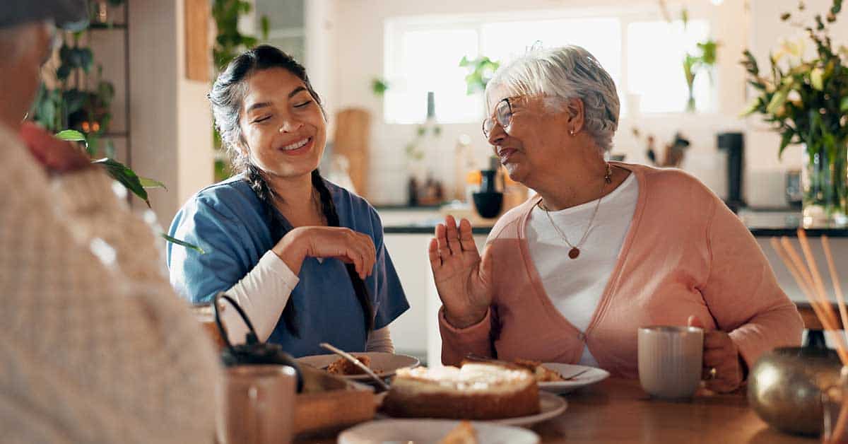 A nurse in scrubs and an older woman sit at a table, smiling and talking over tea and cake in a bright kitchen, demonstrating the importance of safe patient handling while encouraging patient mobility in everyday life.