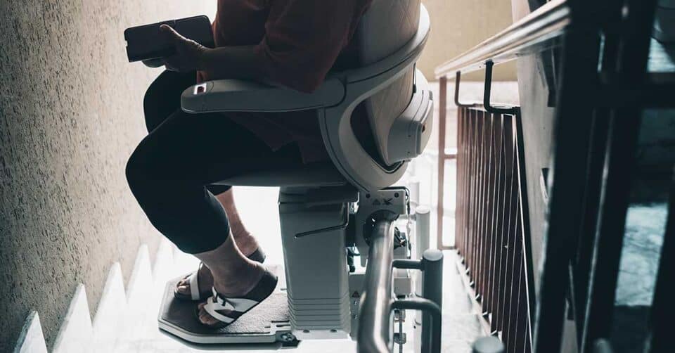 A person sits on a stairlift chair holding a phone, descending a staircase indoors—demonstrating how stairlifts can enhance comfort and accessibility at home.