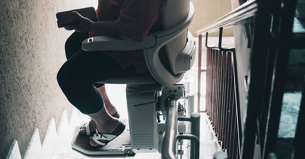 A person sits on a stairlift chair holding a phone, descending a staircase indoors—demonstrating how stairlifts can enhance comfort and accessibility at home.