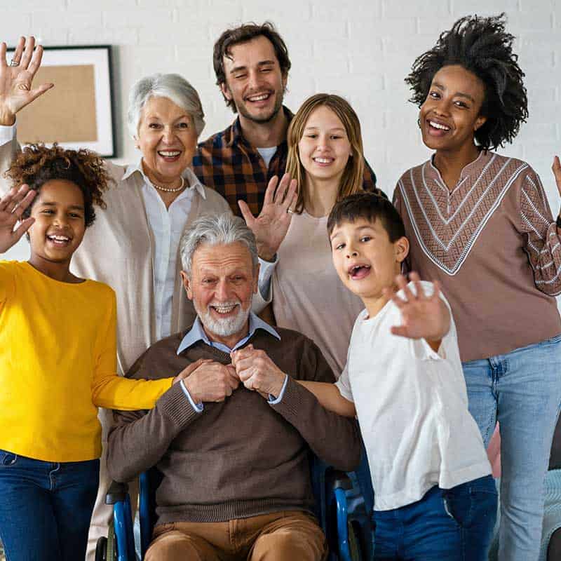 A multigenerational group of people, including children, adults, and seniors, smile and wave at the camera; one man sits in a wheelchair in the front.