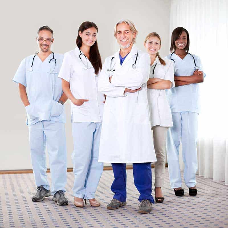 Five healthcare professionals, including doctors and nurses, stand together in a room, wearing medical uniforms and smiling at the camera.