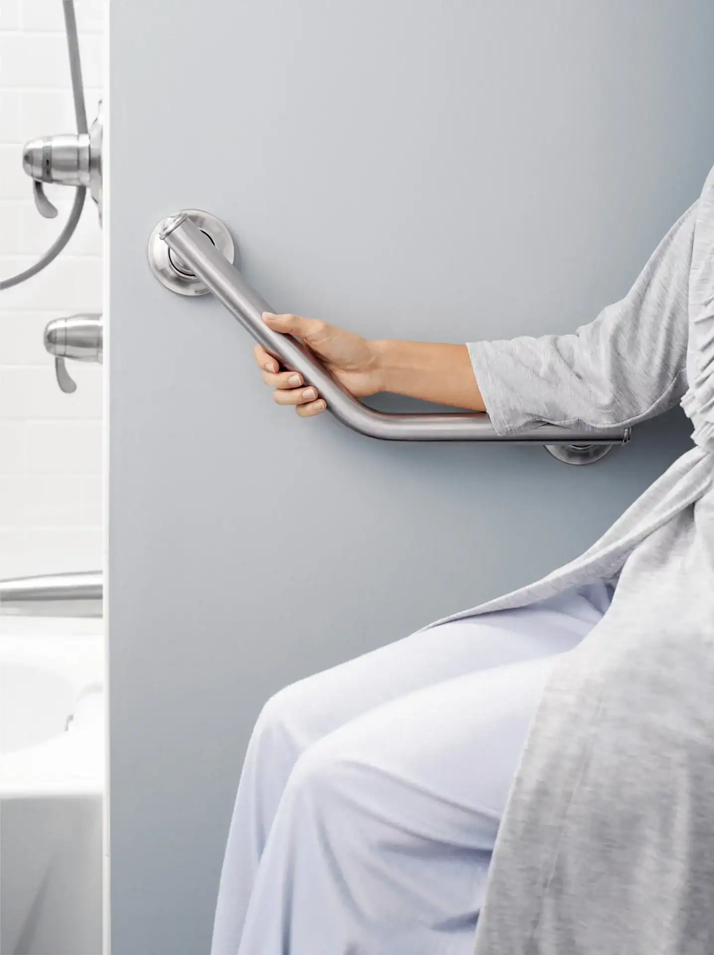 Person sitting on edge of bathtub, holding a silver grab bar—an example of Durable Medical Equipment—attached to a gray wall in a bathroom setting.
