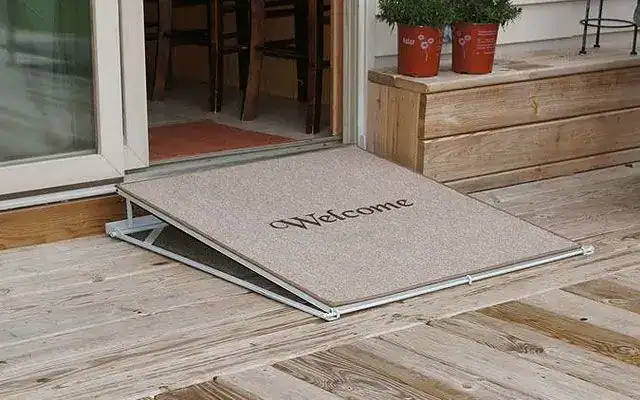 A wooden welcome mat doubles as a ramp, providing wheelchair access to a door on a wooden deck—ideal after an accessibility assessment or consultation services.