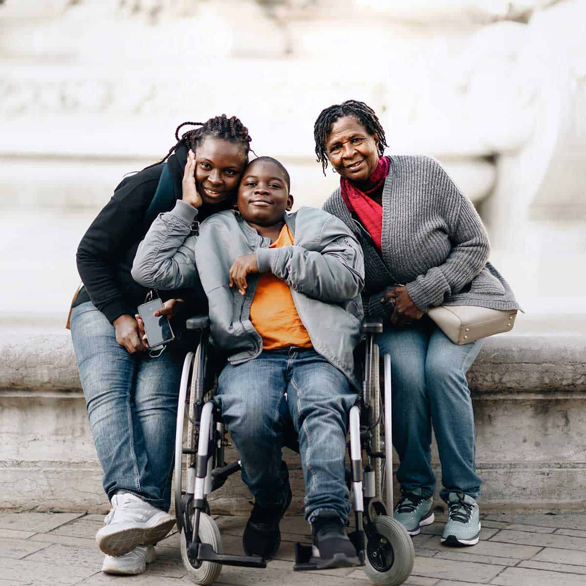 Three people pose for a photo; a boy in a wheelchair is in the center, flanked by two women standing beside him, all smiling outdoors on a stone surface—celebrating the importance of home care for joyful living.