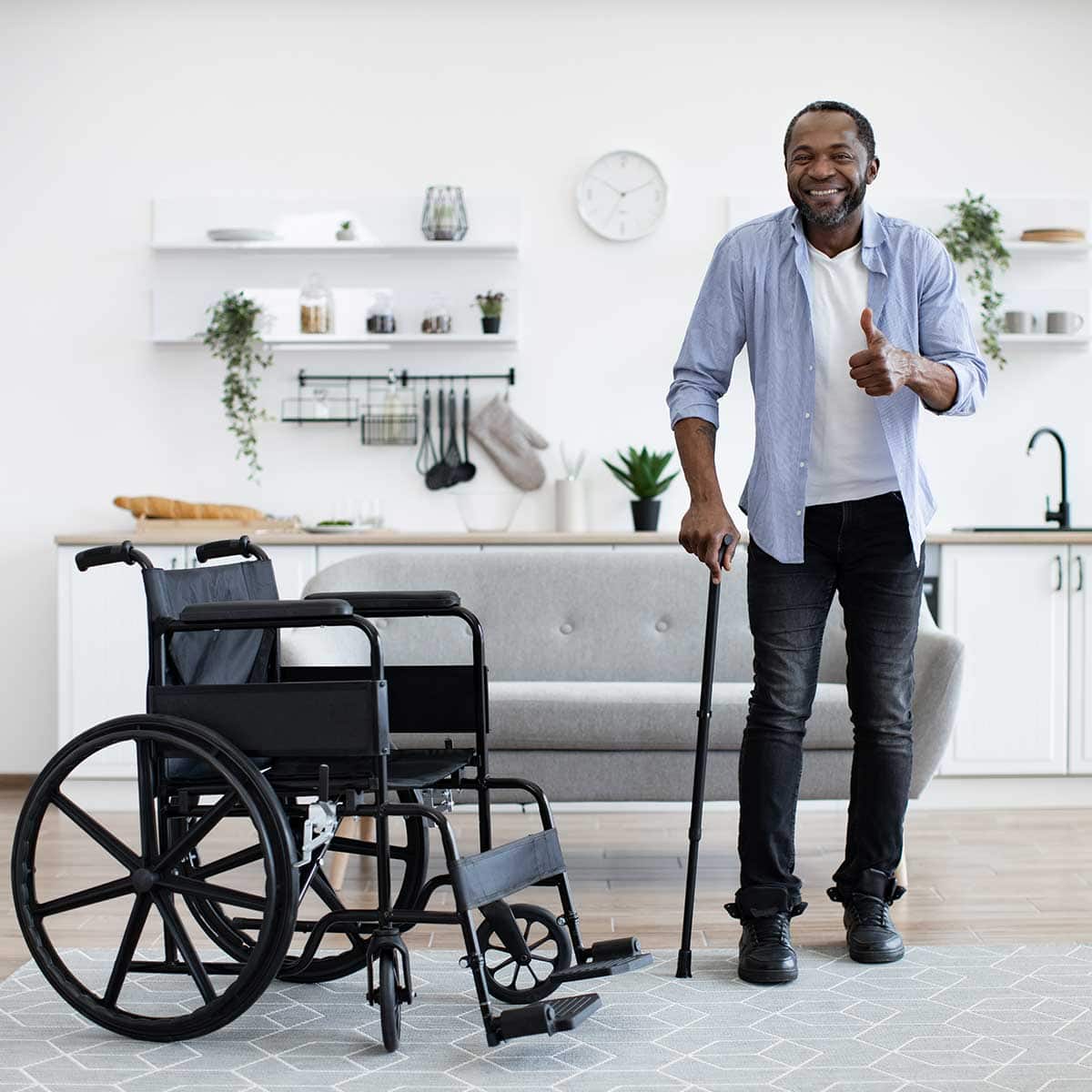 A man standing with a cane gives a thumbs up next to an empty wheelchair in a modern living room, showcasing his Independent Living progress.