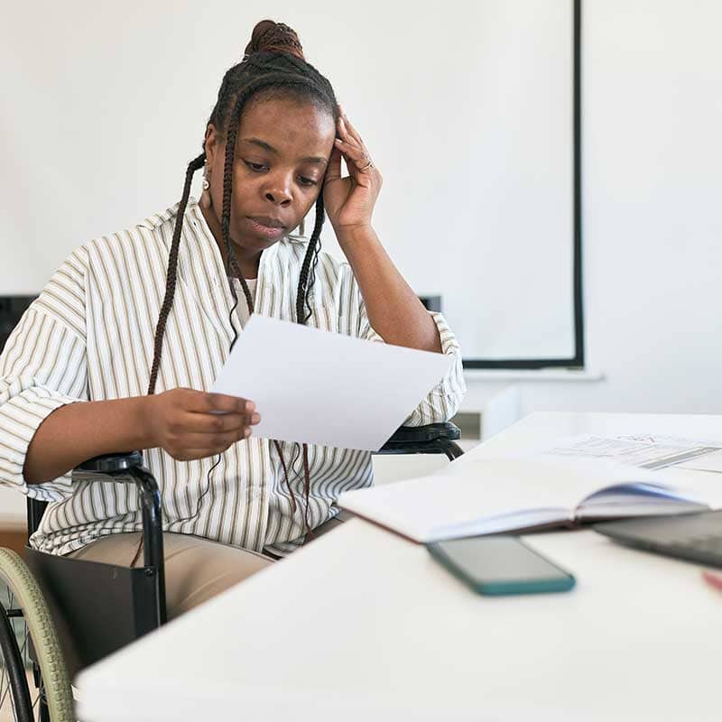 A woman in a wheelchair sits at a desk, reading a document with a concerned expression—perhaps reviewing details related to assessment services. An open notebook and smartphone are on the table beside her.