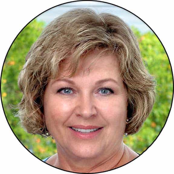 Kristin Hoffman, a woman with short, curly blonde hair and blue eyes, is smiling in an outdoor setting with greenery in the background.