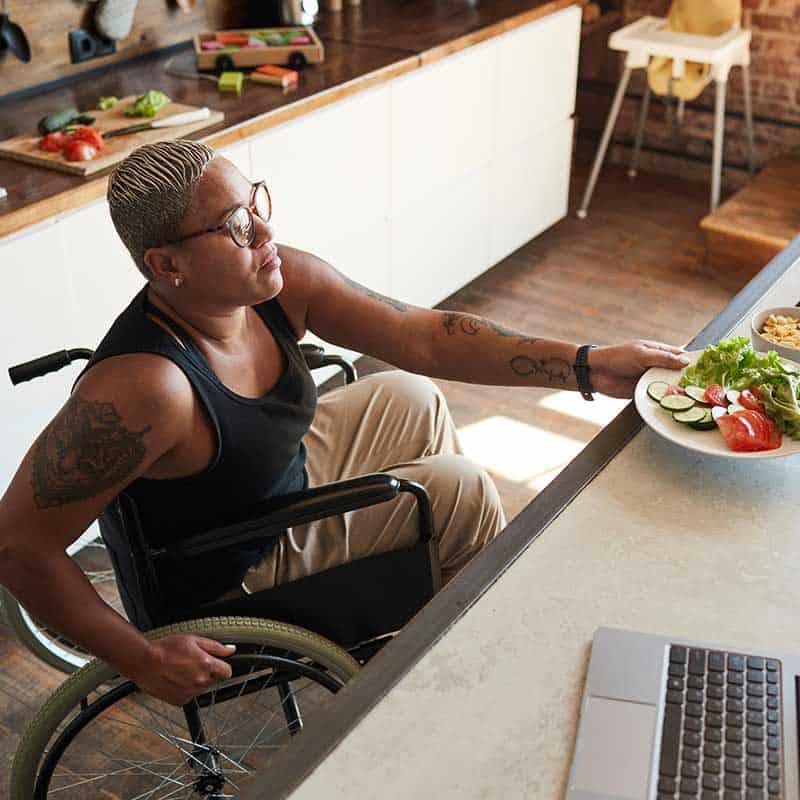 A person in a wheelchair reaches for a plate of fresh vegetables and salad on an accessible kitchen counter, showcasing Home Accessibility Solutions with a laptop and food items nearby.