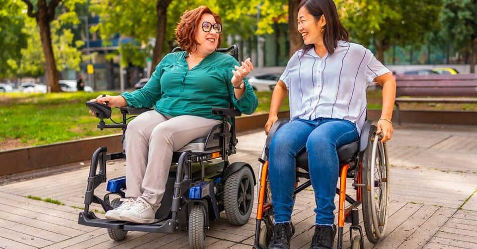 Two women, one in an electric wheelchair and the other in a manual wheelchair, sit outside on a wooden path, talking and smiling together in a park setting.