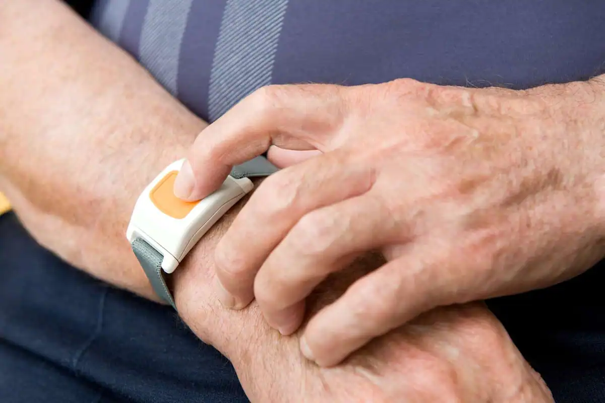 An elderly person presses the button on a medical alert wristband, a piece of durable medical equipment, worn on their left wrist.