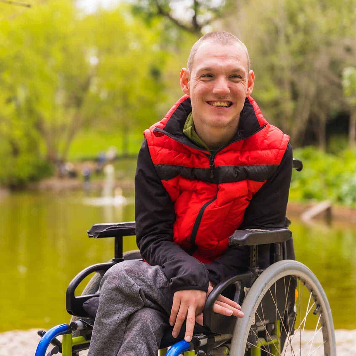 A man in a red vest sits in a wheelchair, a type of durable medical equipment, near a pond in a park, smiling at the camera. Trees and greenery are visible in the background.