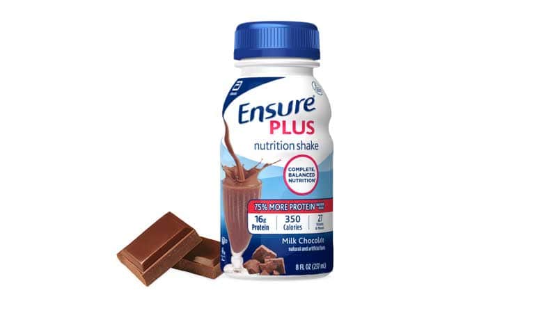 A bottle of Ensure Plus nutrition shake, milk chocolate flavor, is shown next to pieces of chocolate on a white background.
