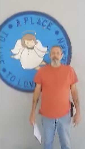 A man in a red shirt stands in front of a wall sign featuring an angel and the words "A Place to Love.