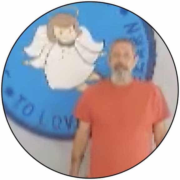 A man in a red shirt stands in front of a blue circular sign featuring an illustrated angel.