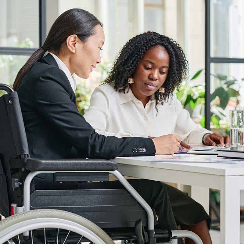 Two women, one in a wheelchair, sit at a table in an office, engaged in an assessment as they review documents together.