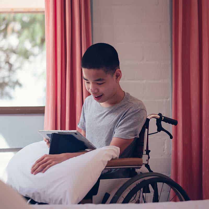 A boy in a wheelchair sits by a window, smiling while using a tablet placed on a pillow on his lap, enjoying the ease provided by Home Accessibility Solutions.