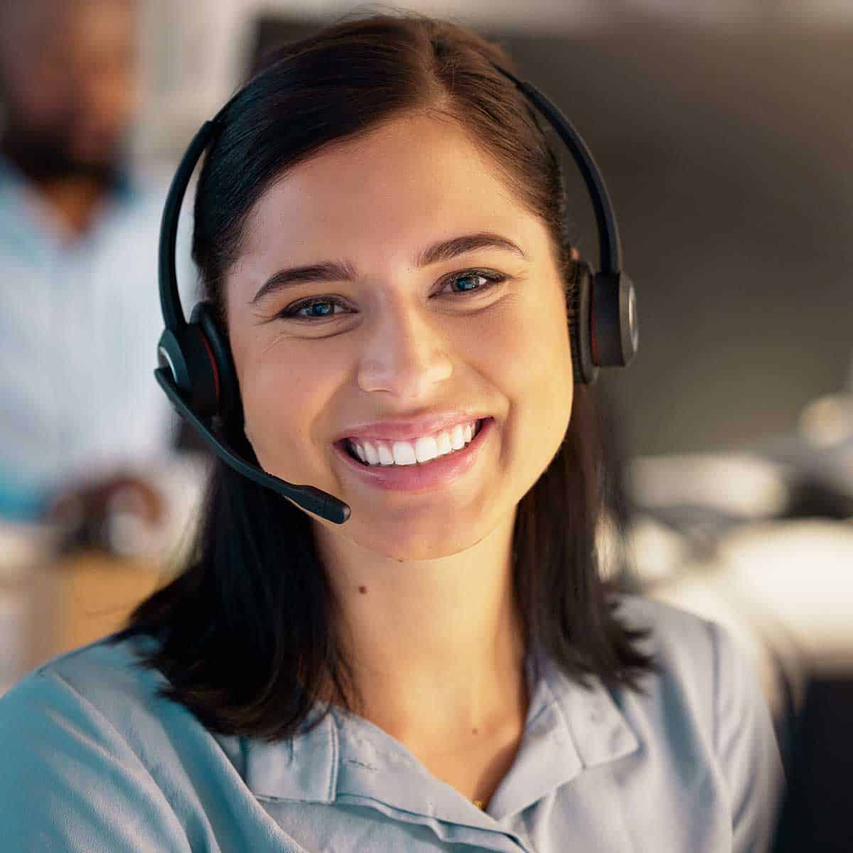 A woman wearing a headset and a light blue shirt smiles while sitting at a desk in an office environment.