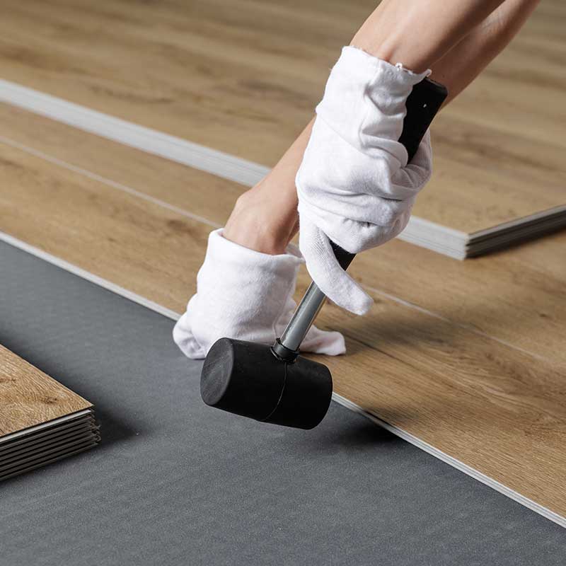 Wearing gloves, a person uses a rubber mallet to install interlocking wooden floor panels on top of an underlayment, creating smooth surfaces that support Home Accessibility Solutions.
