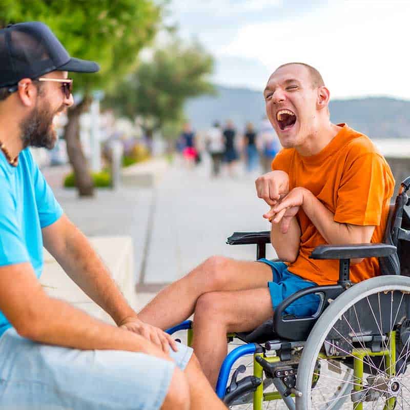 Two men, one in a wheelchair, are outdoors on a sunny day, smiling and laughing together near a walkway with trees and people in the background after receiving consultation services.