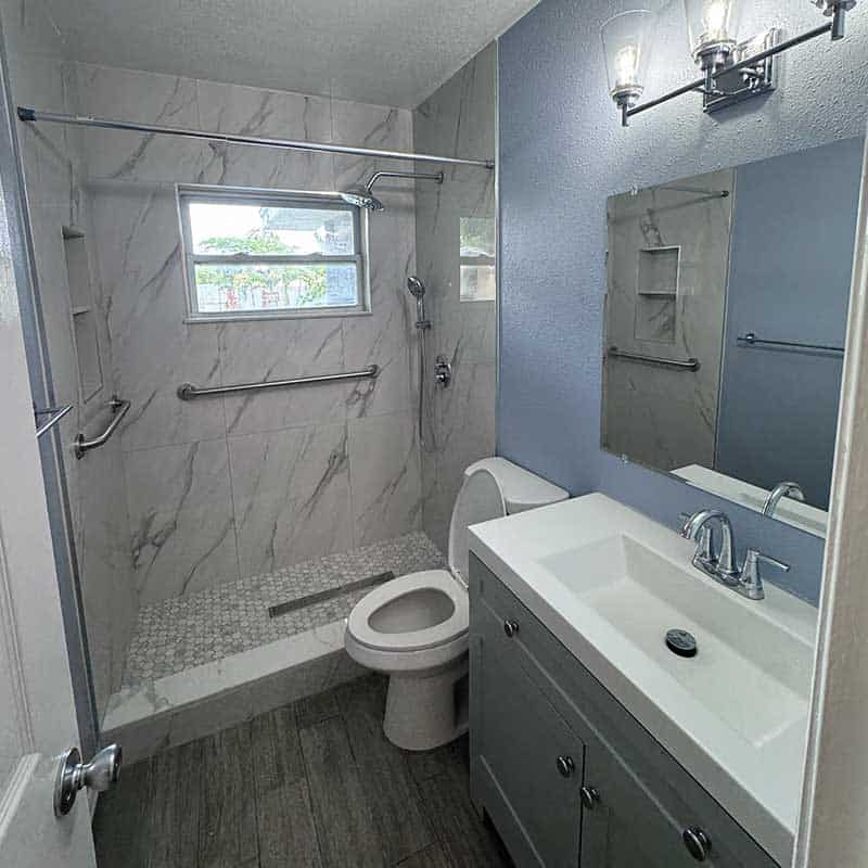 Modern bathroom with a walk-in shower, wall-mounted grab bars, toilet, single-sink vanity, large mirror, overhead lighting, and durable medical equipment for enhanced safety and accessibility.