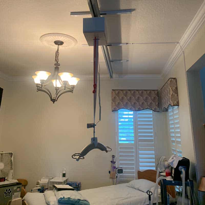 Bedroom with a ceiling-mounted patient lift above a hospital-style bed, medical equipment on tables, and window with shutters and patterned valance—ideal for those seeking Home Accessibility Solutions.