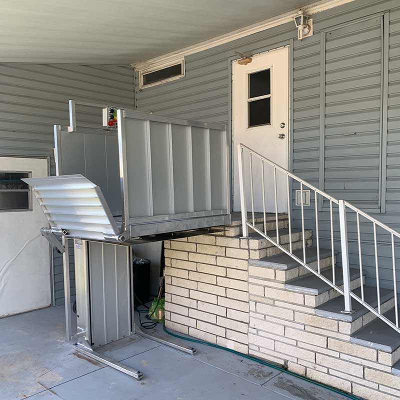 A silver wheelchair lift, part of thoughtful home accessibility solutions, is installed next to a staircase and leads up to a white door on the exterior of a house with gray siding.