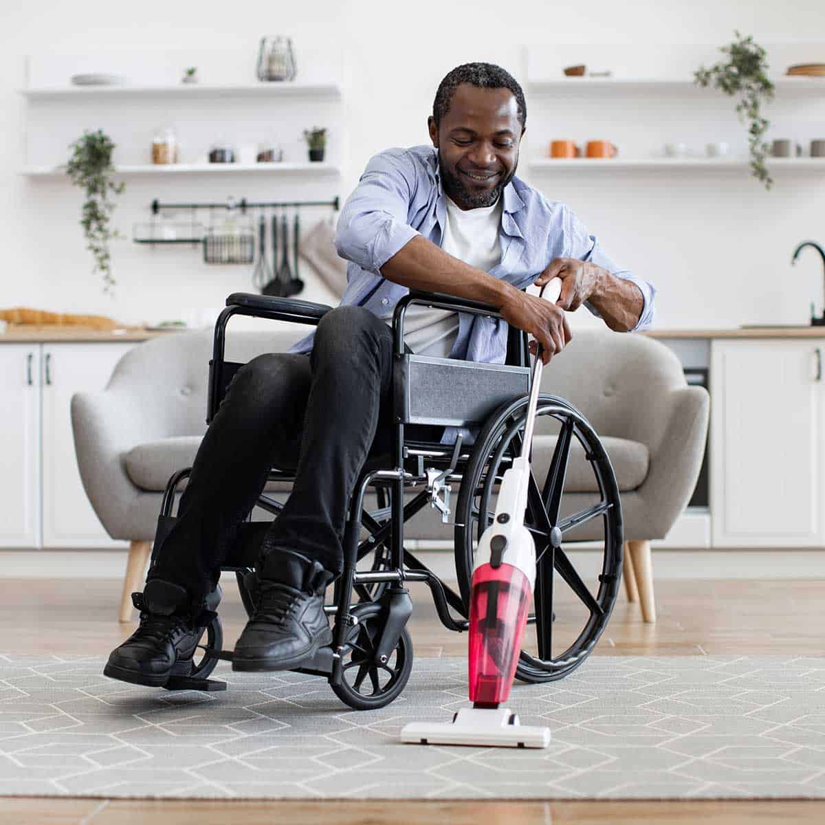 A man in a wheelchair vacuuming a rug in a living room with shelves and a sofa in the background.