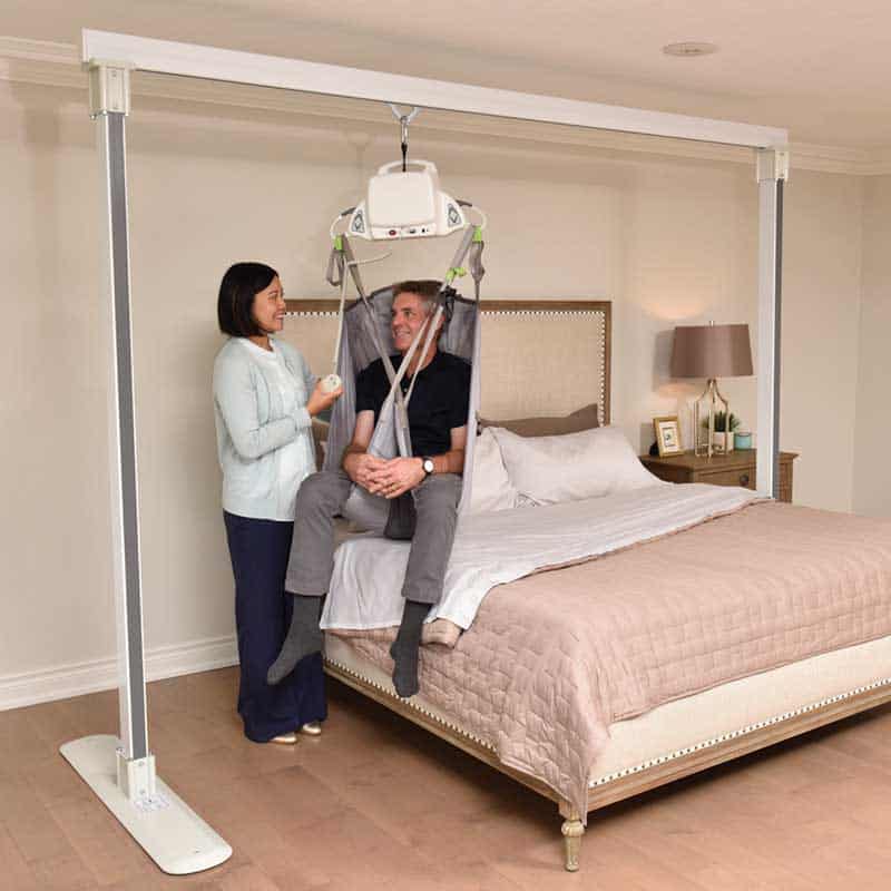 A woman operates a ceiling lift, a type of Durable Medical Equipment, to assist a seated man in a harness over a bed in a bedroom setting.