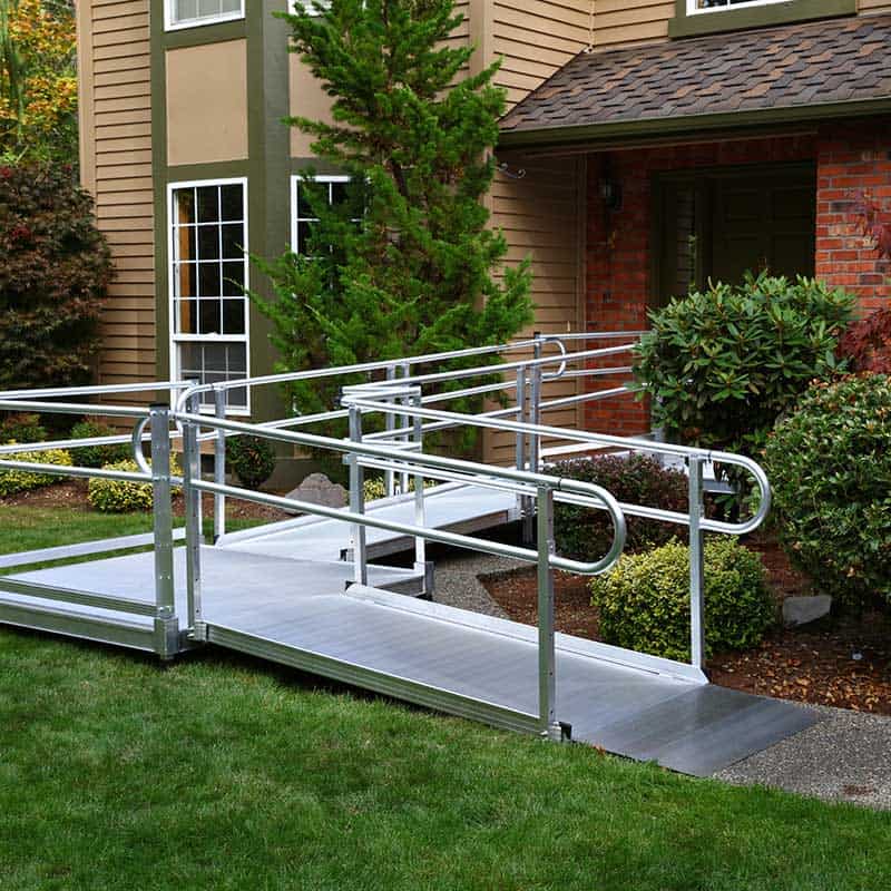 A metal wheelchair ramp with handrails, designed by Home Accessibility Solutions, leads from a sidewalk to the front entry of a house, surrounded by shrubs and grass.